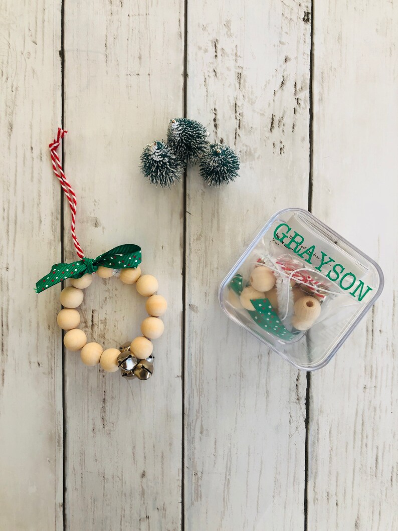 May include: A wooden bead wreath ornament with a green and white polka dot ribbon and a silver bell. The ornament is in a clear plastic container with the name "GRAYSON" written on it. There are also three small green and white pom-poms in the image.