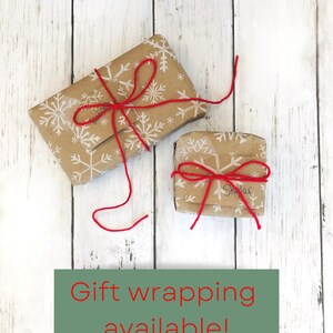 May include: Two small gifts wrapped in brown paper with white snowflakes printed on them. The gifts are tied with red string and bows. The text "Gift wrapping available!" is printed on a green rectangle below the gifts.