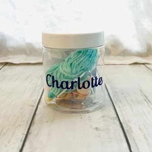 May include: A clear plastic jar with a white lid filled with teal yarn, a gold needle, and a gold bead. The jar has a label that says "Charlotte".