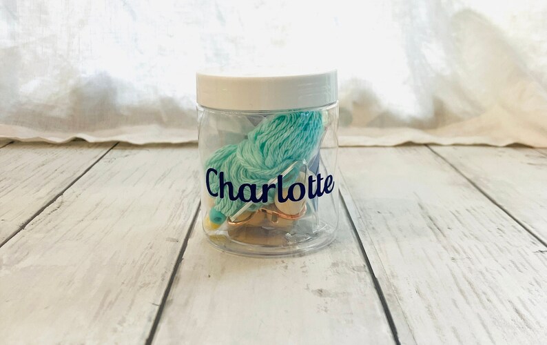 May include: A clear plastic jar with a white lid filled with teal yarn, a gold needle, and a gold bead. The jar has a label that says "Charlotte".