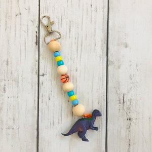 K&ouml;nnte beinhalten: Ein lilafarbener Dinosaurier-Schl&uuml;sselanh&auml;nger mit einer Perlenkette. Die Kette besteht aus Holzperlen, Basketball-Perlen und blauen und gelben Perlen. Der Schl&uuml;sselanh&auml;nger hat einen silbernen Metallverschluss.