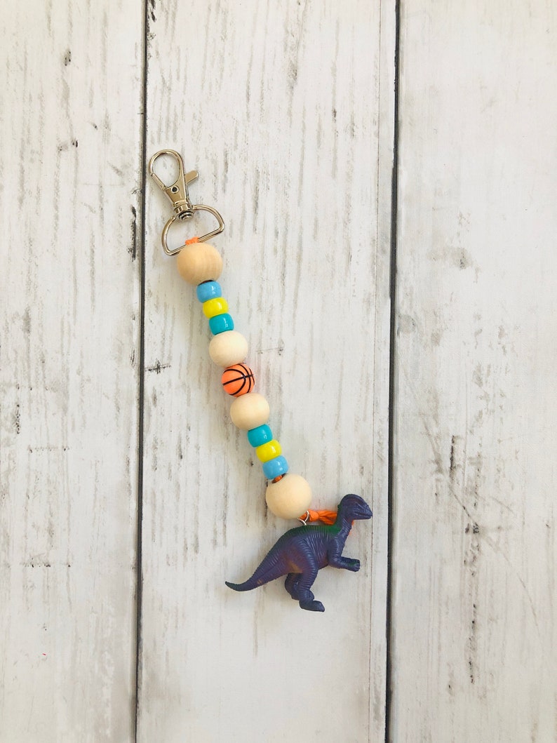 K&ouml;nnte beinhalten: Ein lilafarbener Dinosaurier-Schl&uuml;sselanh&auml;nger mit einer Perlenkette. Die Kette besteht aus Holzperlen, Basketball-Perlen und blauen und gelben Perlen. Der Schl&uuml;sselanh&auml;nger hat einen silbernen Metallverschluss.
