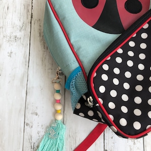 May include: A blue and red ladybug backpack with a black and white polka dot lunch bag. A turquoise tassel keychain with wooden beads hangs from the backpack.