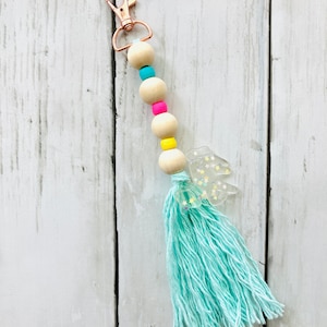 May include: A light blue tassel keychain with a clear charm with gold glitter stars, a gold metal clasp, and a wooden bead chain with pink, yellow, and blue accents.