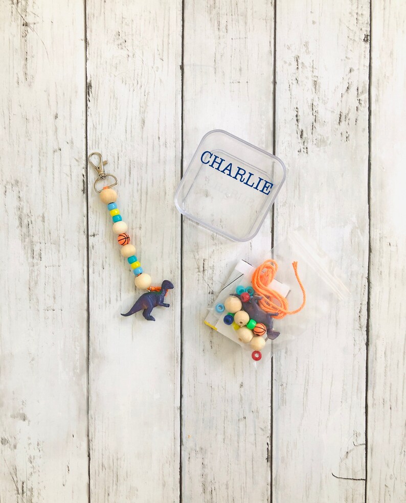 K&ouml;nnte beinhalten: Eine durchsichtige Plastikbox mit dem Namen "CHARLIE" darauf gedruckt. Eine Plastikt&uuml;te enth&auml;lt eine Vielzahl von bunten Perlen, Schnur und ein kleines Dinosaurierspielzeug. Eine Holzperlenkette mit Basketball-Motiven und einem silbernen Schl&uuml;sselanh&auml;nger ist ebenfalls enthalten.