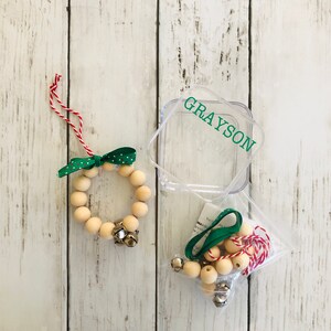May include: A wooden bead wreath ornament kit with a green ribbon, red and white twine, and silver bells. The kit comes in a clear plastic container with the name "GRAYSON" printed on it.