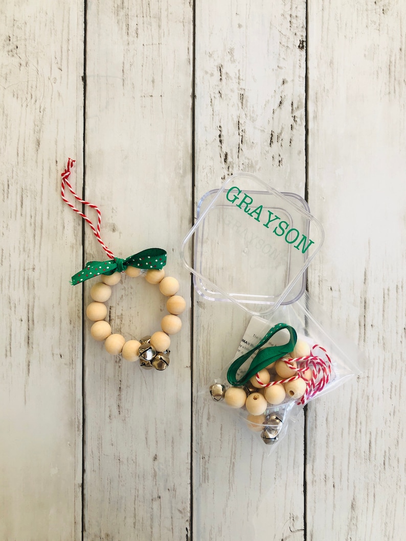 May include: A wooden bead wreath ornament kit with a green ribbon, red and white twine, and silver bells. The kit comes in a clear plastic container with the name "GRAYSON" printed on it.