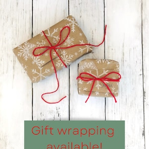 May include: Two small gifts wrapped in brown paper with white snowflakes printed on them. The gifts are tied with red string and bows. The text "Gift wrapping available!" is printed on a green rectangle below the gifts.