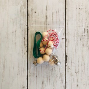 May include: A clear plastic bag filled with craft supplies for making a Christmas ornament. The bag contains wooden beads, jingle bells, green ribbon, and red and white twine.