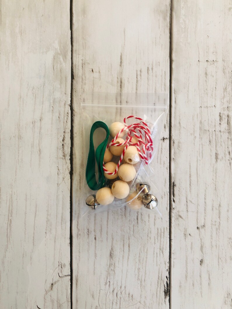 May include: A clear plastic bag filled with craft supplies for making a Christmas ornament. The bag contains wooden beads, jingle bells, green ribbon, and red and white twine.