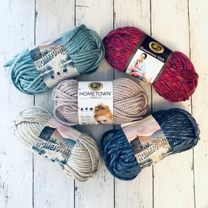 Pode incluir: Cinco novelos de fio em diferentes cores, incluindo azul, cinza, bege e vermelho. O fio é etiquetado como "Hometown" e "Hometown USA".