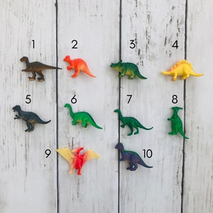 K&ouml;nnte beinhalten: Ein Set aus zehn bunten Plastik-Dinosaurier-Spielzeugen, die von 1 bis 10 nummeriert sind. Die Dinosaurier sind in zwei Reihen auf einem wei&szlig;en Holzgrund angeordnet. Die Dinosaurier sind alle in verschiedenen Farben und Gr&ouml;&szlig;en.