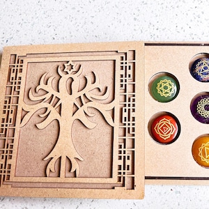 May include: Wooden box with a tree of life design on the front and seven round stones with chakra symbols inside. The stones are in different colours, including red, orange, yellow, green, blue, indigo, and violet.
