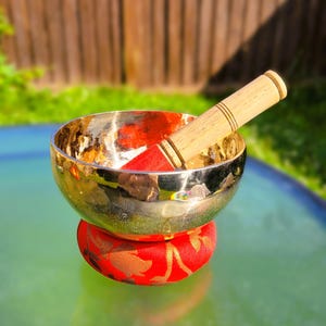 Puede incluir: Un cuenco cant&oacute;n dorado con un mazo de madera, descansando sobre un coj&iacute;n rojo con dise&ntilde;os florales dorados. El cuenco est&aacute; sobre una superficie verde.