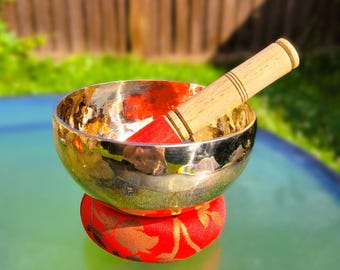 May include: A gold singing bowl with a wooden striker, resting on a red cushion with gold floral designs. The bowl is sitting on a green surface.