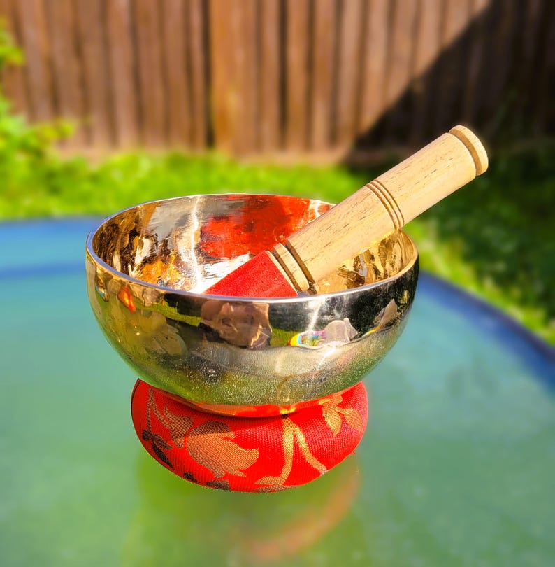 Puede incluir: Un cuenco cant&oacute;n dorado con un mazo de madera, descansando sobre un coj&iacute;n rojo con dise&ntilde;os florales dorados. El cuenco est&aacute; sobre una superficie verde.