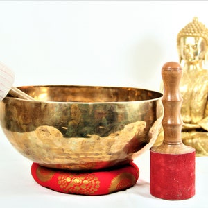 Puede incluir: Un cuenco cantor dorado con un mazo de madera y un coj&iacute;n rojo. Una estatua dorada de Buda est&aacute; en el fondo.