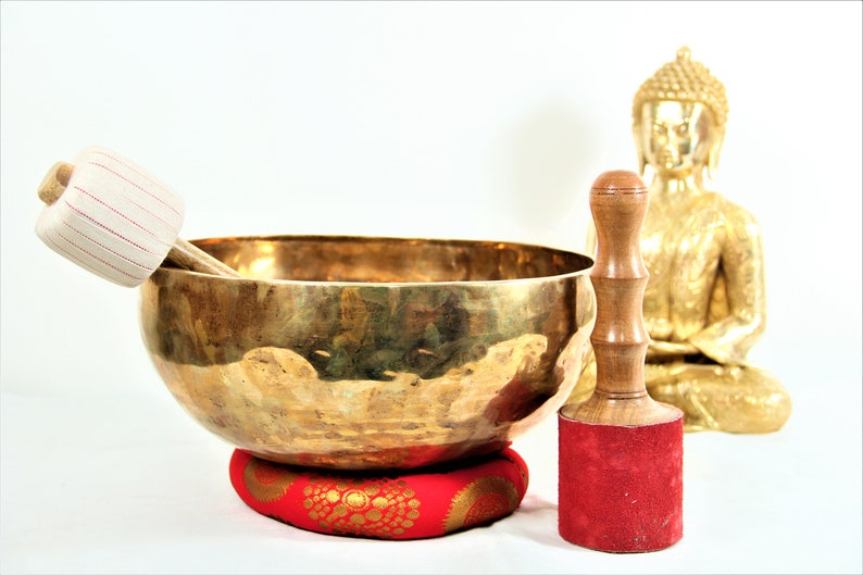 Puede incluir: Un cuenco cantor dorado con un mazo de madera y un coj&iacute;n rojo. Una estatua dorada de Buda est&aacute; en el fondo.