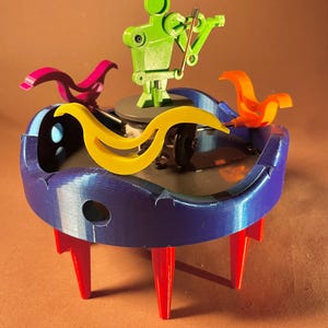 May include: A whimsical, colorful sculpture featuring a green robot playing a violin. The sculpture has a blue base with red legs, yellow and pink curved accents, and an orange bird-like figure. The design is modern and playful.
