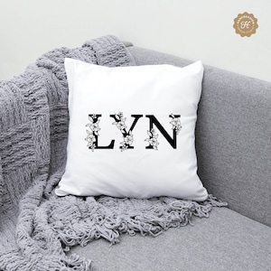K&ouml;nnte beinhalten: Ein wei&szlig;es Kissen mit dem Namen "LYN" in schwarzen Buchstaben mit floralen Akzenten.