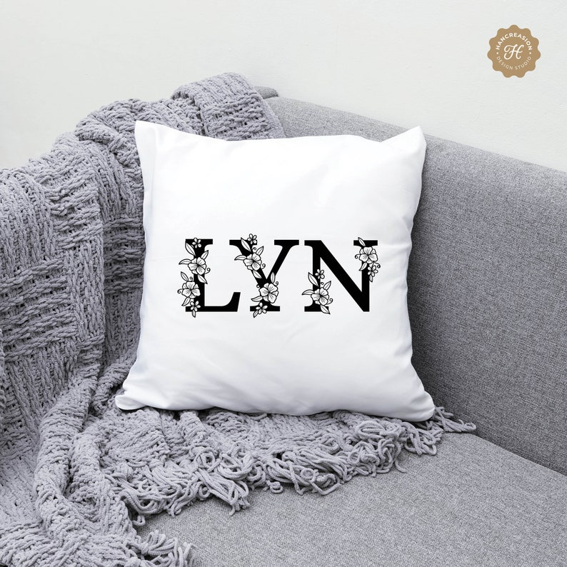 K&ouml;nnte beinhalten: Ein wei&szlig;es Kissen mit dem Namen "LYN" in schwarzen Buchstaben mit floralen Akzenten.