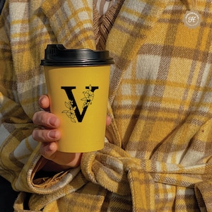 K&ouml;nnte beinhalten: Ein gelber To-Go-Becher mit schwarzem Deckel und einem schwarzen floralen Design mit dem Buchstaben V in der Mitte.