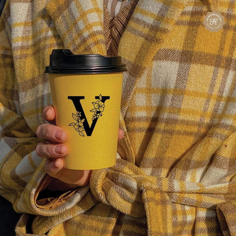 K&ouml;nnte beinhalten: Ein gelber To-Go-Becher mit schwarzem Deckel und einem schwarzen floralen Design mit dem Buchstaben V in der Mitte.