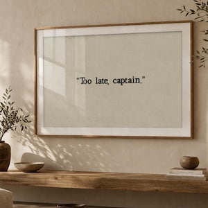 May include: A framed print with the text "Too late, captain." hangs above a wooden shelf. The shelf displays decorative bowls, books, and a mushroom-shaped lamp. A vase with leafy branches and a sofa are also visible.