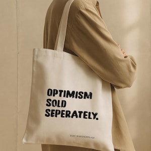 Op de afbeelding: Een witte draagtas met zwarte tekst die luidt "OPTIMISM SOLD SEPERATELY". De tas heeft een rechthoekige vorm met een handvat en de tekst "TINY MANIFESTO #58" onderaan. De tas wordt door een persoon vastgehouden.