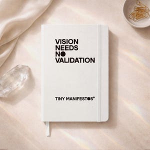 Op de afbeelding: Een wit notitieboek met de tekst "VISION NEEDS NO VALIDATION" en "TINY MANIFESTOS*". Het notitieboek heeft een wit lint-bladwijzer. Gouden paperclips liggen in een kleine witte kom. Een heldere kristal is ook zichtbaar.