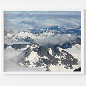 May include: An aerial view of a snow-capped mountain range with clouds swirling around the peaks. The mountains are rugged and jagged, with some areas covered in snow and ice. The sky is a light blue, and the clouds are white and fluffy.