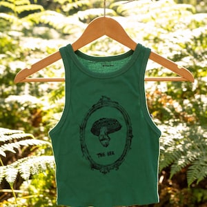 Peut inclure: Débardeur vert avec un motif de champignon noir dans un cadre ovale. Le texte "THE BEE" est en dessous. Le débardeur est suspendu à un cintre en bois, sur un fond vert flou.