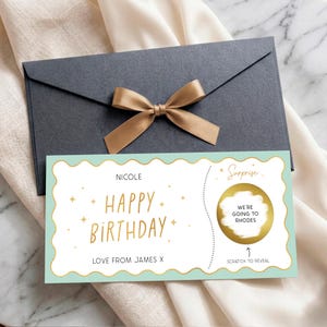 May include: A dark grey envelope with a gold ribbon bow and a rectangular scratch-off birthday card. The card features a light green scalloped border, gold text reading "HAPPY BIRTHDAY" and "NICOLE", and a gold circle with "WE'RE GOING TO RHODES".