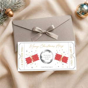 Könnte beinhalten: Ein rechteckiges weißes Geschenkticket mit schwarzem Rand, goldenem Text Merry Christmas Amy und A GIFT FOR YOU an den Seiten. Es zeigt rote gemusterte Knallbonbons, goldene Sterne und einen silbernen Rubbelkreis mit der Aufschrift We are going to see Moulin Rouge!.