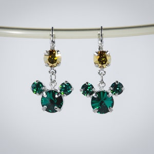 May include: A pair of silver dangle earrings with green and gold crystal accents. The earrings feature a cluster of three green crystals in the shape of a Mickey Mouse head.