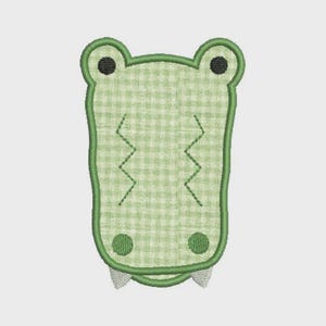 Puede incluir: Cabeza de cocodrilo bordada con un patrón de cuadros verdes y blancos. El cocodrilo tiene ojos negros, detalles en zigzag y dos dientes pequeños. El contorno y los detalles son verdes.