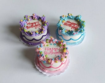 Può includere: Tre torte rotonde in miniatura con glassa bianca, decorate con bordi colorati, zuccherini e la scritta Happy Birthday in corsivo rosso o rosa. Le torte presentano accenti viola, blu e rosa sui lati e sulla parte superiore.