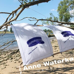 Könnte beinhalten: Zwei weiße Stoffsegel mit blauem Hutdesign und dem Text "Ahoi Hamburg!" werden an einem Sandstrand präsentiert. Die Segel sind an Holzrahmen befestigt. Das Bild enthält auch den Text "Anne Waterkant HAMBURG".
