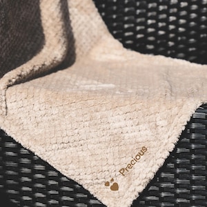 May include: A beige plush blanket with the word "Precious" embroidered on it. The blanket is folded and placed on a black wicker chair.