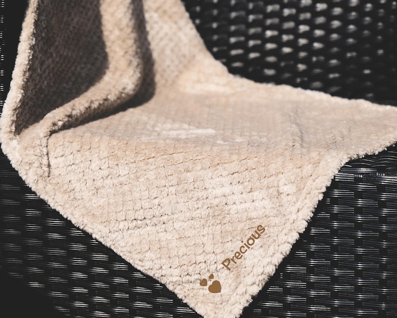 May include: A beige plush blanket with the word "Precious" embroidered on it. The blanket is folded and placed on a black wicker chair.
