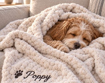 May include: A cream-colored, chunky knit throw blanket featuring a soft, textured surface. The blanket is embroidered with a dark brown paw print icon and the name Poppy in a cursive script. A small, light brown dog is sleeping under the blanket.