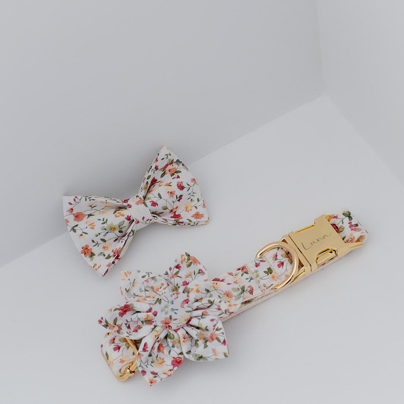 May include: A white floral dog collar with a gold buckle and a matching bow tie. The collar has the word "Luna" engraved on the buckle.