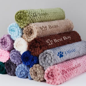 May include: A stack of plush blankets in various colors, each with a personalized name embroidered on them. The names include Sandy, Beau, Best Boy, Olive, and Gandalf.