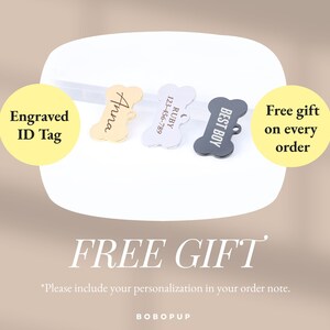 May include: Three dog bone-shaped engraved ID tags. The tags are gold, silver, and black. The gold tag says "Anna", the silver tag says "RUBY 123-456-789", and the black tag says "BEST BOY". The text "Engraved ID Tag" is in a yellow circle on the left side of the image. The text "Free gift on every order" is in a yellow circle on the right side of the image. The text "FREE GIFT" is in large letters below the tags. The text "*Please include your personalization in your order note." is below the large text. The text "BOBOPUP" is at the bottom of the image.