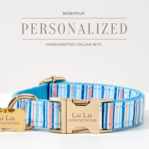 May include: A personalized dog collar set with a blue, pink, and white striped pattern. The set includes a matching collar and bone-shaped tag with the name "Lu Lu" and a phone number. The collar has a gold-colored buckle and hardware. The text "PERSONALIZED HANDCRAFTED COLLAR SETS" is displayed.