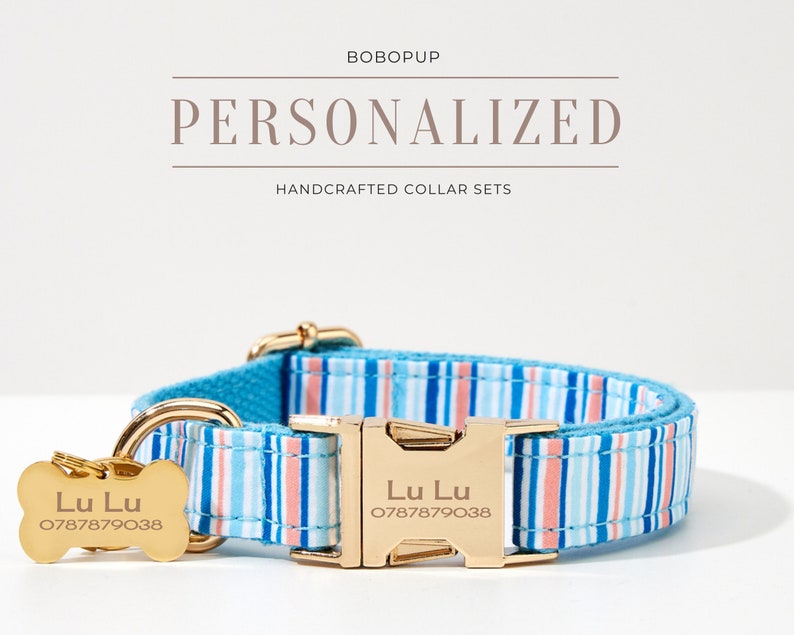 May include: A personalized dog collar set with a blue, pink, and white striped pattern. The set includes a matching collar and bone-shaped tag with the name "Lu Lu" and a phone number. The collar has a gold-colored buckle and hardware. The text "PERSONALIZED HANDCRAFTED COLLAR SETS" is displayed.