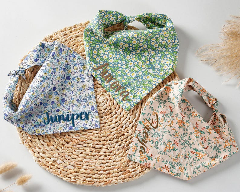May include: Three floral patterned dog bandanas in blue, green, and peach, each with a name embroidered on them. The bandanas are arranged on a round woven mat. The blue bandana has the name "Juniper", the green bandana has the name "Aurora", and the peach bandana has the name "Theia".