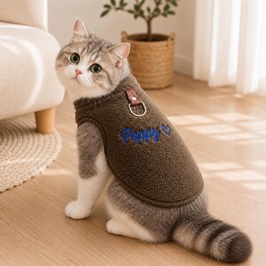 May include: A brown fleece pet vest featuring a textured sherpa-like material, a silver metal D-ring attached with a brown faux leather tab at the neckline, and the name Poppy embroidered in blue script with a small blue heart beside it.