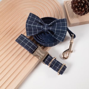 Puede incluir: Un juego de collar y correa para perro de cuadros azul marino y blanco con hebilla dorada y pajarita a juego. El collar tiene una etiqueta personalizada que dice "Mowgli 159.248.7890".