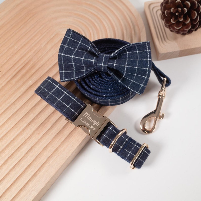 Puede incluir: Un juego de collar y correa para perro de cuadros azul marino y blanco con hebilla dorada y pajarita a juego. El collar tiene una etiqueta personalizada que dice "Mowgli 159.248.7890".